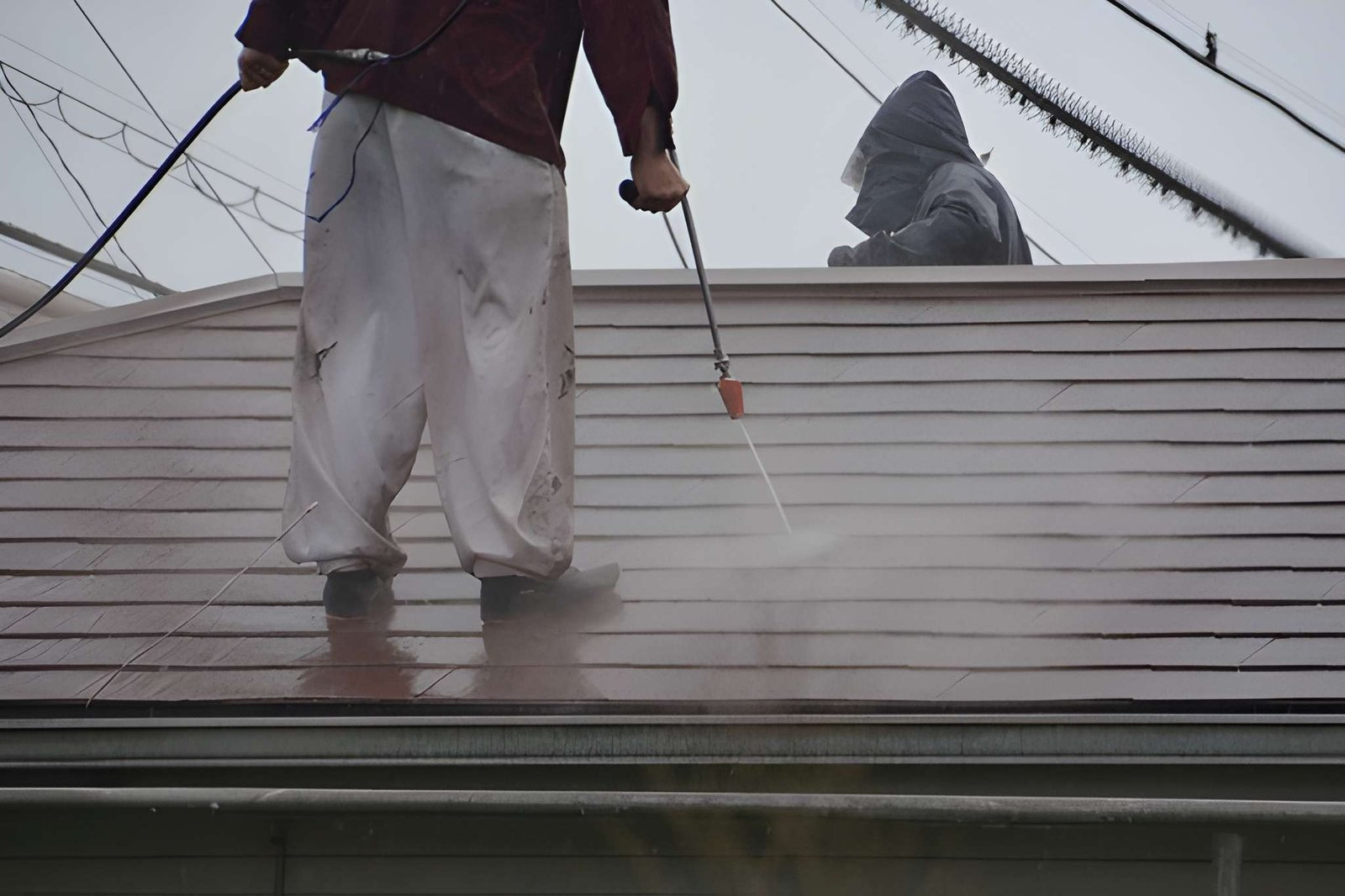 A professional cleaner standing on a sloped residential roof, using a high-pressure power washer wand to clean dirt and moss off brown roof tiles.