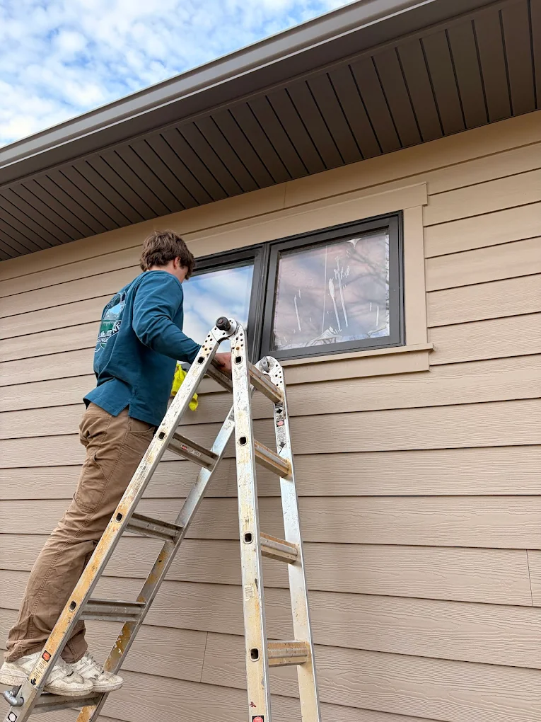 A technician standing on an A-frame extension ladder cleaning a high residential window with a microfiber cloth on a tan-sided house.